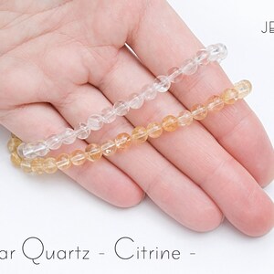May include: Two beaded bracelets, one clear quartz and one citrine, rest on a hand. The clear quartz bracelet is transparent, while the citrine bracelet has a warm, golden-yellow hue. The image includes the text "- Clear Quartz - Citrine -" in a handwritten font.