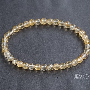 May include: A bracelet made of small, round, translucent, amber-colored beads. The beads are strung together on a clear elastic band. The bracelet is displayed on a dark gray surface. The word "JEWOSS" is printed in white.