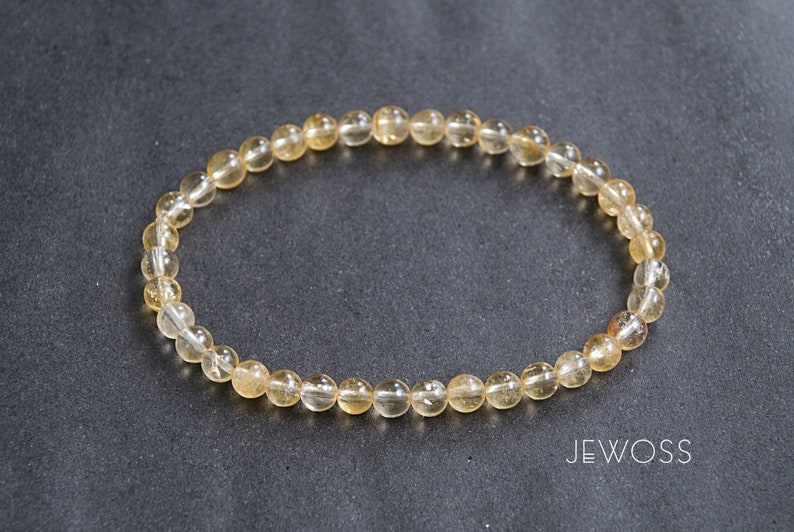 May include: A bracelet made of small, round, translucent, amber-colored beads. The beads are strung together on a clear elastic band. The bracelet is displayed on a dark gray surface. The word "JEWOSS" is printed in white.