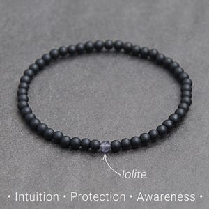 Può includere: Braccialetto di perline nere con una singola, piccola pietra di iolite blu-viola. Il braccialetto è composto da piccole perline nere opache. Il testo in basso recita: Intuizione, Protezione, Consapevolezza.