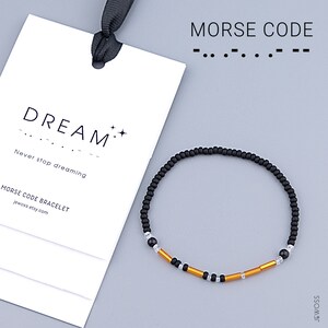 Puede incluir: Una pulsera de cuentas negras con detalles dorados y transparentes. La pulsera está junto a una etiqueta blanca con la palabra "DREAM" y el texto "Never stop dreaming". Las palabras "MORSE CODE" están en la parte superior de la imagen.