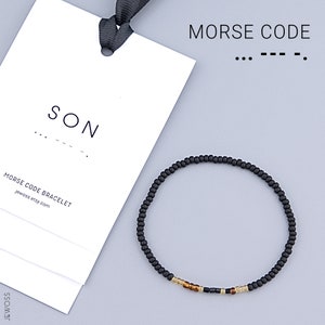 May include: Black beaded bracelet with gold and black beads spelling out "SON" in Morse code. The bracelet is on a white card with the text "MORSE CODE BRACELET jewoss.etsy.com".