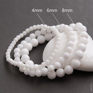 Puede incluir: Tres hebras de cuentas redondas blancas, cada hebra con un tamaño de cuenta diferente: 4 mm, 6 mm y 8 mm. Las cuentas están dispuestas en un círculo sobre una piedra blanca.