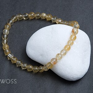 May include: A beaded bracelet composed of small, translucent, golden-yellow gemstones. The bracelet is resting on a white, rounded stone against a dark gray background. The brand name "JEWOSS" is visible in the lower left corner.