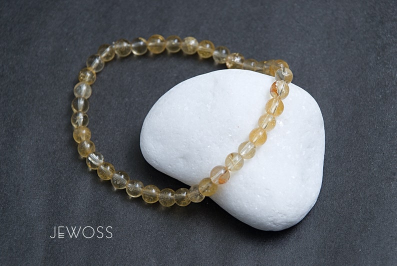 May include: A beaded bracelet composed of small, translucent, golden-yellow gemstones. The bracelet is resting on a white, rounded stone against a dark gray background. The brand name "JEWOSS" is visible in the lower left corner.