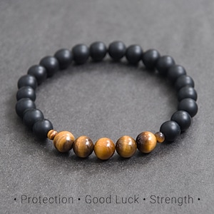 May include: A beaded bracelet featuring matte black onyx beads and tiger's eye beads. The bracelet is strung on an elastic cord. The bracelet is displayed on a gray surface. The text at the bottom says "Protection • Good Luck • Strength."