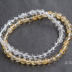 May include: Two beaded bracelets. One is clear, the other is a golden yellow. The beads are small and round. The bracelets are lying on a dark gray surface. The brand name "JEWOSS" is visible in white text.