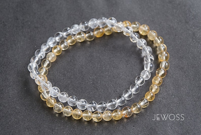 May include: Two beaded bracelets. One is clear, the other is a golden yellow. The beads are small and round. The bracelets are lying on a dark gray surface. The brand name "JEWOSS" is visible in white text.
