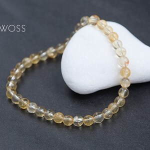 May include: A beaded bracelet with golden-yellow, translucent beads. The bracelet is resting on a white, rounded stone against a dark gray background. The brand name "JEWOSS" is in white text in the upper left corner.