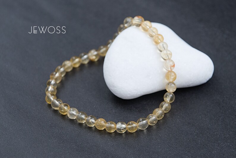 May include: A beaded bracelet with golden-yellow, translucent beads. The bracelet is resting on a white, rounded stone against a dark gray background. The brand name "JEWOSS" is in white text in the upper left corner.