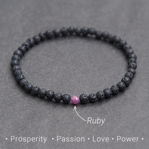 May include: A black lava stone bracelet with a single pink ruby bead. The bracelet is on a gray surface. The text "Ruby" is written below the bracelet. The text "Prosperity, Passion, Love, Power" is written below the text "Ruby".