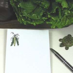 May include: A white notepad with a green pea pod illustration. The notepad is on a white surface with a pen and a small turtle figurine.