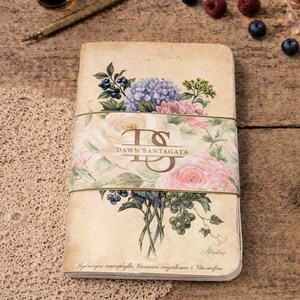 May include: A vintage-style notebook with floral illustrations, including roses, hydrangeas, and berries. The cover features the text "DAWN SANTAGATA" and is bound with a gold elastic band. An ink bottle and pen are on the wooden surface.