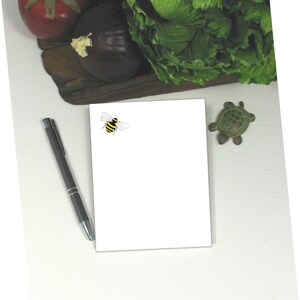 May include: A white notepad with a small bee illustration in the upper left corner. A pen and a small turtle figurine are on the white surface.