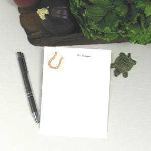 May include: A white notepad with a brown baseball illustration and the text "Dawn Sundagala" at the top. A silver pen and a small green turtle figurine are on the surface.