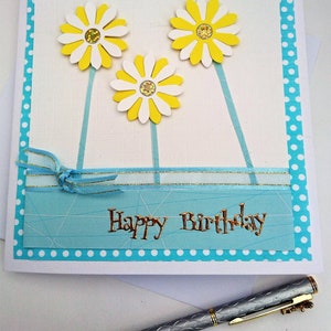 Op de afbeelding: Handgemaakte verjaardagskaart met drie gele en witte madeliefjes. De kaart heeft een blauwe stippelrand en de woorden "Happy Birthday" in goud. Een zilveren pen met gouden accenten ligt eronder.