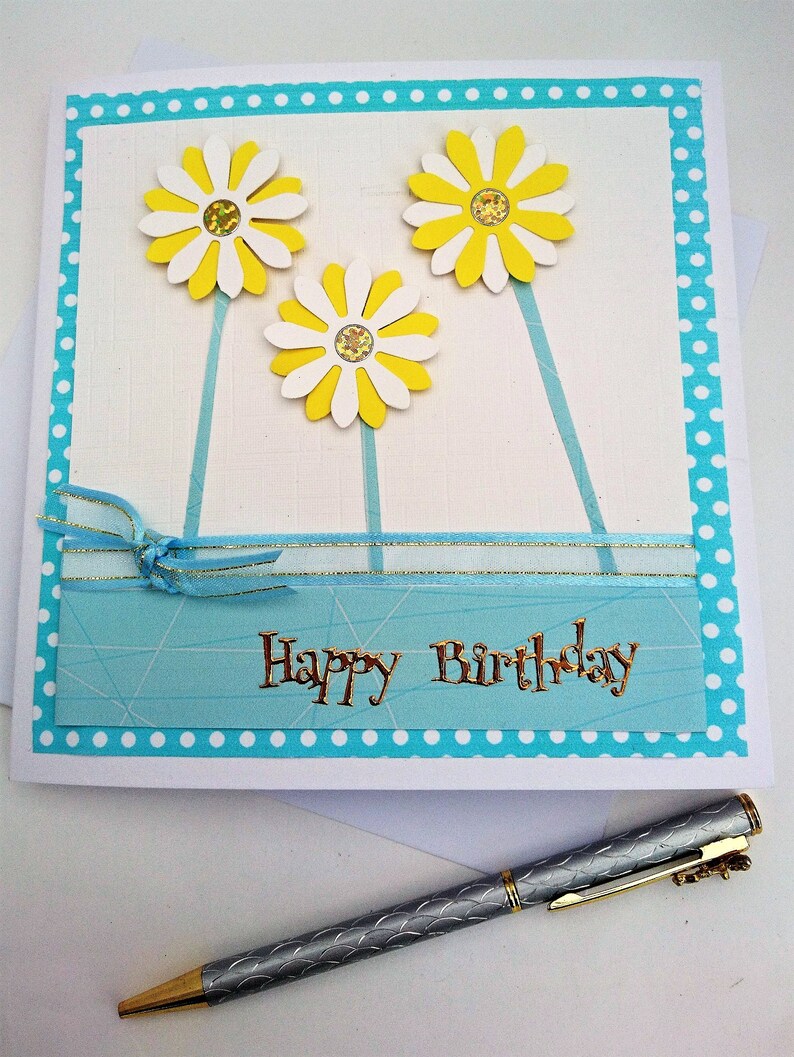 Op de afbeelding: Handgemaakte verjaardagskaart met drie gele en witte madeliefjes. De kaart heeft een blauwe stippelrand en de woorden "Happy Birthday" in goud. Een zilveren pen met gouden accenten ligt eronder.