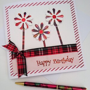 Op de afbeelding: Een handgemaakte verjaardagskaart met drie witte en rode madeliefachtige bloemen. De kaart heeft een rood geruit lint en de tekst "Happy Birthday". Een bijpassende geruite pen ligt eronder.