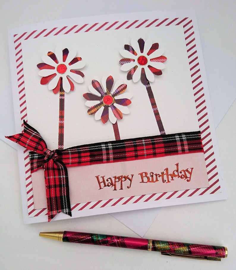 Op de afbeelding: Een handgemaakte verjaardagskaart met drie witte en rode madeliefachtige bloemen. De kaart heeft een rood geruit lint en de tekst "Happy Birthday". Een bijpassende geruite pen ligt eronder.