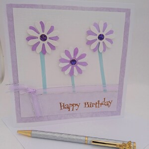 Op de afbeelding: Een handgemaakte verjaardagskaart met drie witte en paarse madeliefbloemen, elk met een paarse edelsteen in het midden. De kaart heeft een lichtpaarse rand en de woorden "Happy Birthday" in goud. Een zilveren pen met gouden details ligt ervoor.