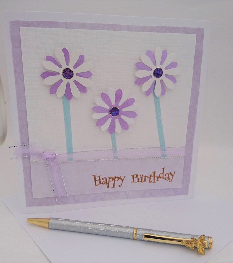 Op de afbeelding: Een handgemaakte verjaardagskaart met drie witte en paarse madeliefbloemen, elk met een paarse edelsteen in het midden. De kaart heeft een lichtpaarse rand en de woorden "Happy Birthday" in goud. Een zilveren pen met gouden details ligt ervoor.