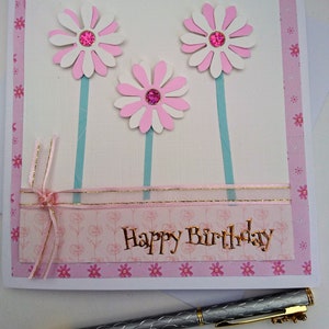 Op de afbeelding: Handgemaakte verjaardagskaart met drie witte en roze madeliefbloemen, elk met een roze edelsteencentrum. De kaart heeft een roze bloemenrand en gouden letters met de tekst "Happy Birthday". Een zilveren pen ligt op de voorgrond.