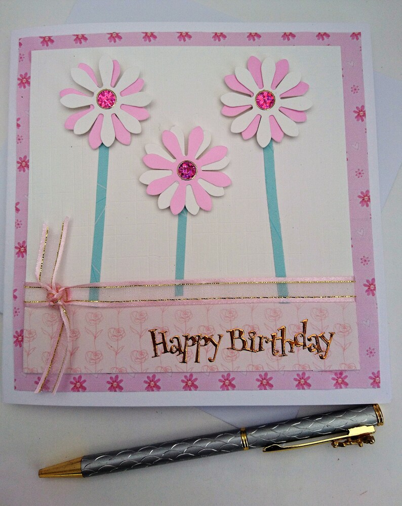 Op de afbeelding: Handgemaakte verjaardagskaart met drie witte en roze madeliefbloemen, elk met een roze edelsteencentrum. De kaart heeft een roze bloemenrand en gouden letters met de tekst "Happy Birthday". Een zilveren pen ligt op de voorgrond.