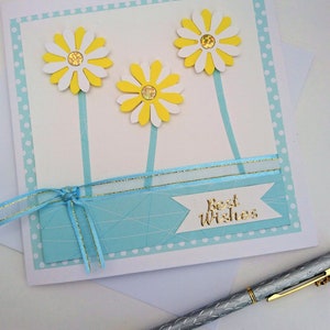 Op de afbeelding: Handgemaakte wenskaart met drie gele en witte madeliefjes. De kaart heeft een lichtblauwe achtergrond met een lint en de woorden "Best Wishes" in goud. Een zilveren pen met een gouden punt is zichtbaar.