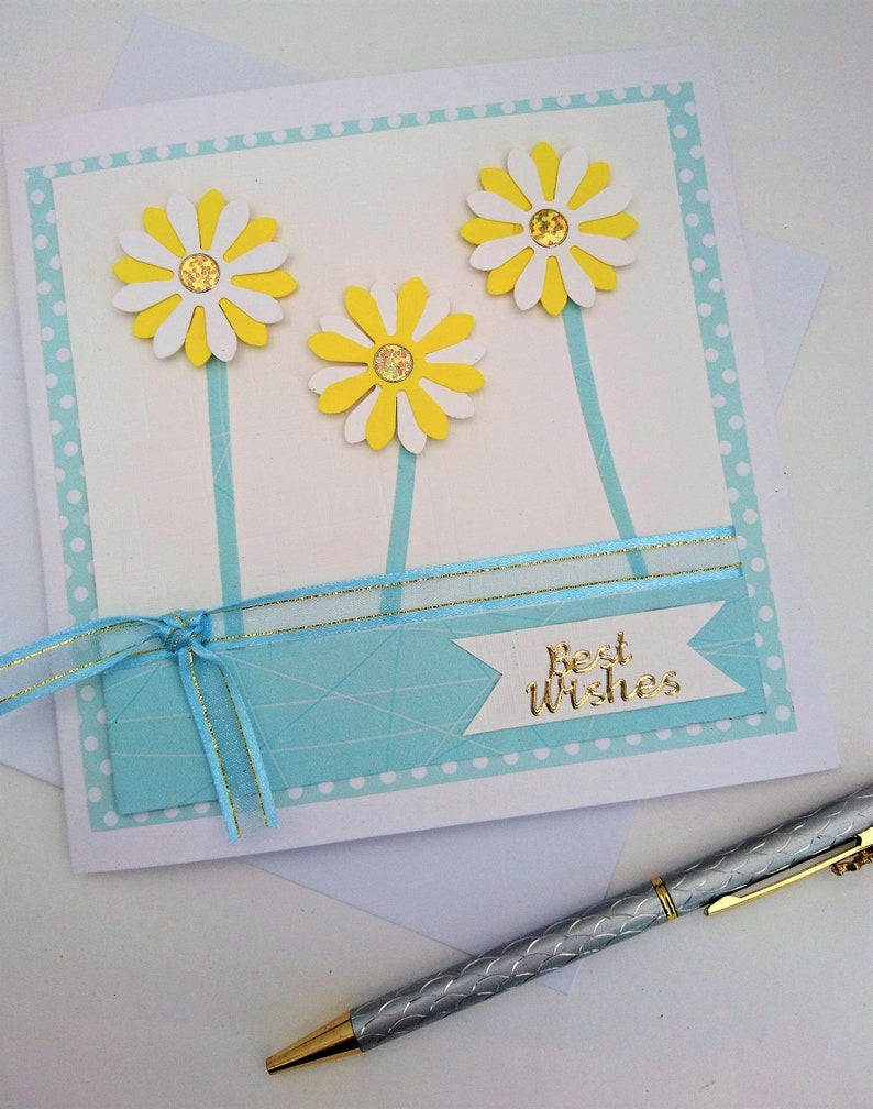 Op de afbeelding: Handgemaakte wenskaart met drie gele en witte madeliefjes. De kaart heeft een lichtblauwe achtergrond met een lint en de woorden "Best Wishes" in goud. Een zilveren pen met een gouden punt is zichtbaar.