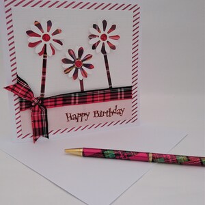 Op de afbeelding: Een verjaardagskaart met drie bloemversieringen en een bijpassende pen. De kaart heeft een rood-zwarte geruite lint en de woorden "Happy Birthday". De pen heeft een gouden punt en een ruitpatroon.