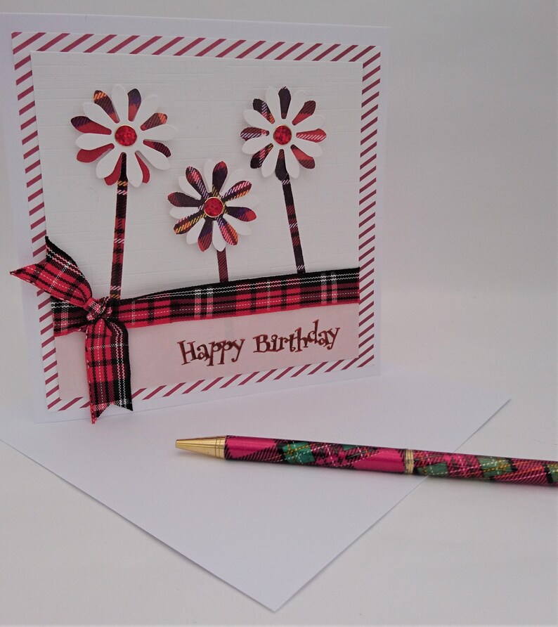 Op de afbeelding: Een verjaardagskaart met drie bloemversieringen en een bijpassende pen. De kaart heeft een rood-zwarte geruite lint en de woorden "Happy Birthday". De pen heeft een gouden punt en een ruitpatroon.
