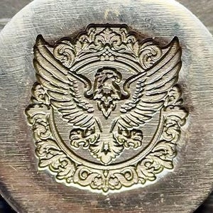May include: A circular metal stamp featuring a deeply engraved design of an eagle with outstretched wings, centered within an ornate, scrolling filigree border. The metallic surface shows a brushed texture, highlighting the intricate relief details.