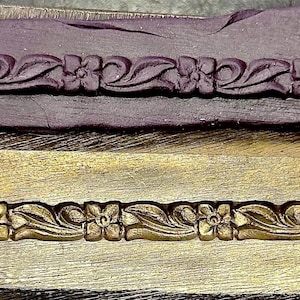 Peut inclure: Deux blocs d'impression en bois. Le bloc supérieur est violet foncé et rectangulaire avec un motif floral sculpté le long du bord inférieur. Le bloc inférieur est de couleur or avec un motif floral similaire.