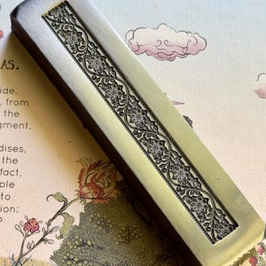 May include: A rectangular metal paperweight with a brushed silver finish and a central recessed panel featuring an intricate, repeating floral vine pattern. The object has a dark, solid base and a smooth, metallic top surface with detailed engraving.