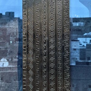 May include: A rectangular, silver-colored metal object with a repeating Greek key pattern. The design is embossed, creating a textured surface. The object is likely a decorative element or part of a larger piece.