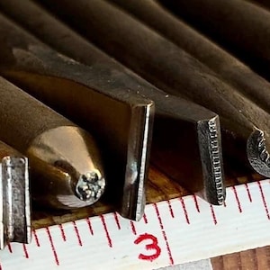May include: A collection of metal leather stamping tools with various shaped tips, including flat, curved, and textured designs, arranged side-by-side next to a white measuring tape showing the tools are between 7.6 and 10.2 cm in length.