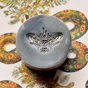 May include: A circular, silver metal stamp featuring a detailed embossed moth design. The stamp is set against a backdrop of colourful, patterned artwork with snakes and floral elements. The moth design is intricate.