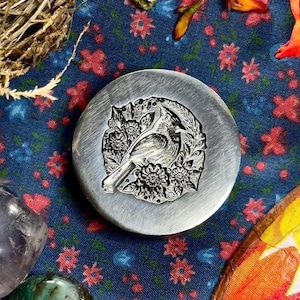May include: A circular silver-toned metal wax seal stamp featuring an embossed design of a cardinal bird perched among detailed flowers and leaves. The stamp has a brushed metallic finish and a clear, intricate relief of the bird and floral motif.