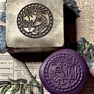 May include: A square metal stamp featuring a circular engraved design of a flute player, a sun, and a cactus, paired with a round purple wax seal impression showing the same detailed motif of the flute player, sun, and cactus in relief.