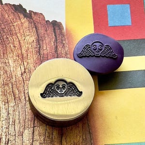 May include: Two wax seal stamps featuring a skull and wing design. One is gold-coloured, the other purple. The stamps are set against a colourful background with a wood-grain texture.