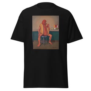 Puede incluir: Una camiseta negra de manga corta con un estampado gráfico de una persona disfrazada de perrito caliente sentada en un taburete. El personaje sostiene una botella de condimento roja, con varias botellas en un mostrador al fondo.