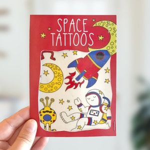 May include: A red and white package with the text "SPACE TATTOOS" on the front. The package contains temporary tattoos of an astronaut, a rocket, a moon, and a space alien.