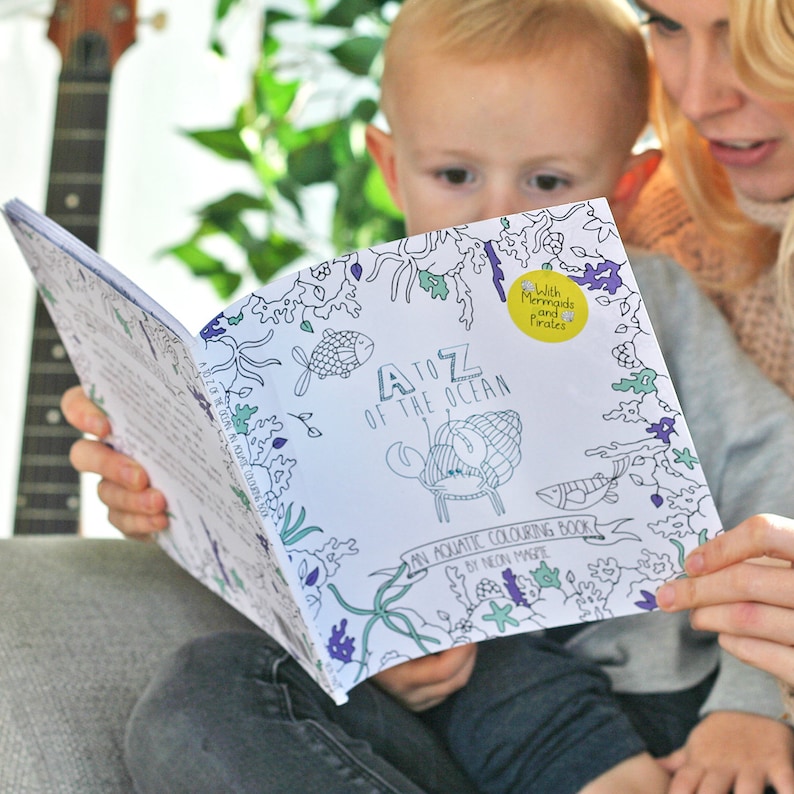 May include: A child is looking at a coloring book titled "A to Z of the Ocean: An Aquatic Coloring Book by Neon Magpie". The book has a colorful illustration of a crab and seaweed on the cover. A sticker with the text "With Mermaids and Pirates" is on the cover.