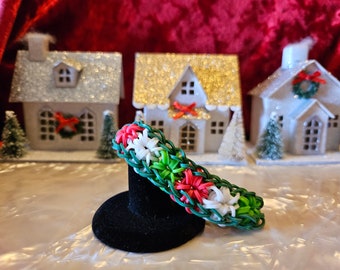 May include: A green, white and red rubber band bracelet with a flower design. The bracelet is on a black display stand.