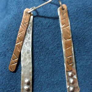 May include: A pair of silver and copper earrings with a hammered texture. The earrings are long and narrow, with a silver base and a copper strip on top. The copper strip has a geometric pattern and the silver base has three silver beads at the bottom.