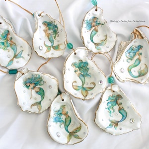 May include: A collection of seven oyster shell ornaments, each featuring a hand-painted mermaid with flowing blue and green hair and a matching tail. The shells are accented with small clear rhinestones, gold-toned edges, and gold hanging cords with teal beads. The text Cathy's Colorful Creations is visible.