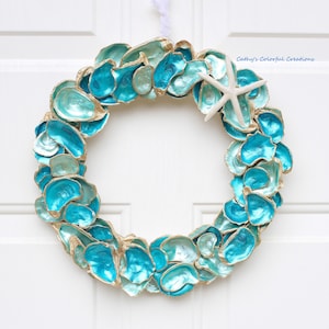 May include: A circular wreath constructed from numerous oyster shells painted in varying shades of turquoise, teal, and blue with gold-toned edges. A single white starfish is attached to the upper right side. The text Cathy's Colorful Creations is visible.
