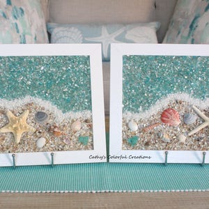 May include: Two framed beach art pieces with white frames. Each features a sandy beach scene with seashells, starfish, and turquoise glass representing the ocean. The art is displayed on a teal placemat.