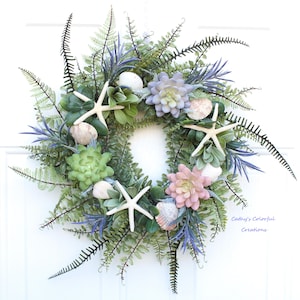 May include: A decorative wreath featuring a variety of faux greenery, including ferns and succulents, with seashells and white starfish accents. The wreath has a circular shape and is set against a white background. The text "Cathy's Colorful Creations" is visible.