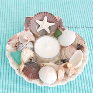 May include: A seashell candle holder with a white candle in the center. The holder is decorated with various seashells, a white starfish, and small pieces of sea glass. The background is a light blue fabric.